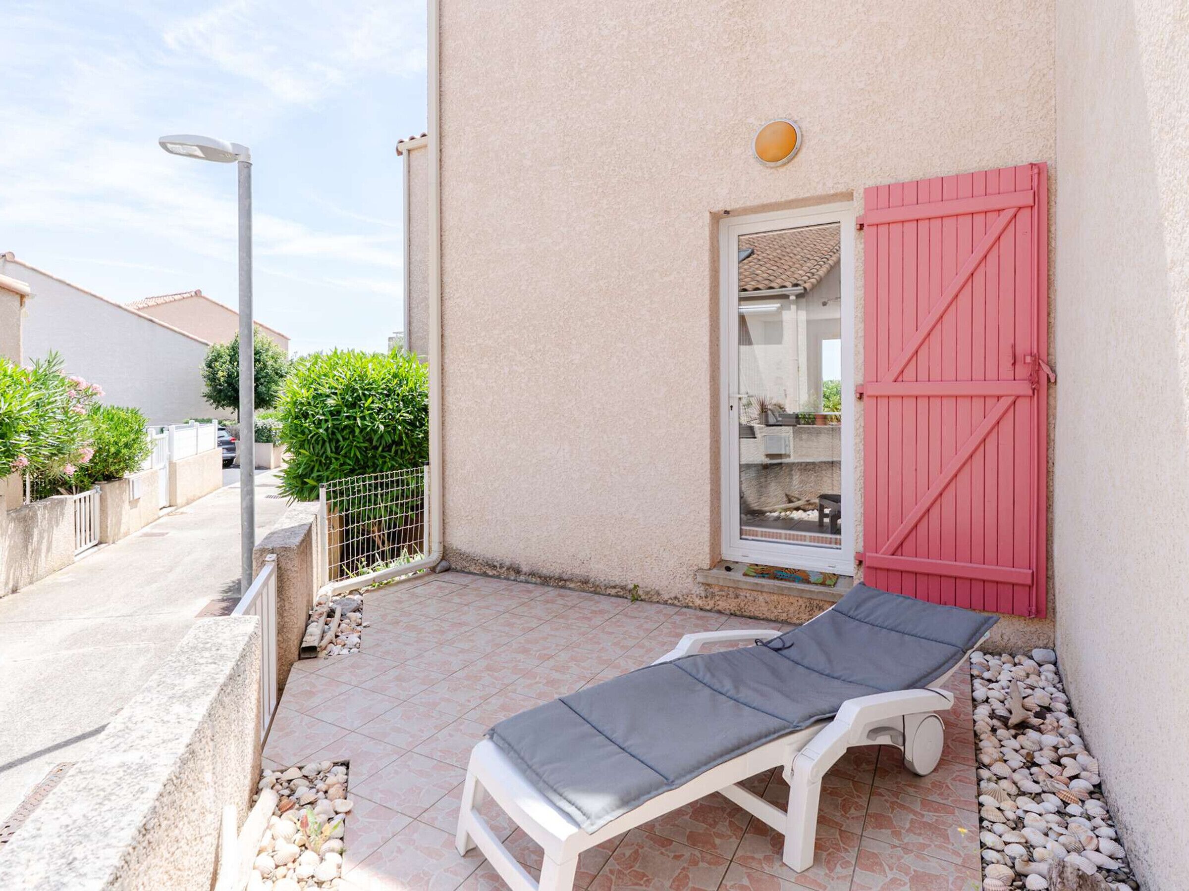Un espacio exterior iluminado por el sol, con una silla de lounge blanca, una puerta con contraventanas rojas y una ventana pequeña.