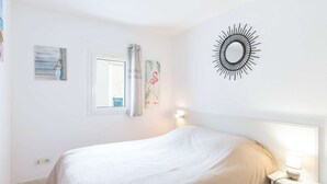 A bedroom with a bed, a nightstand with a lamp, a window with curtains, and wall art. - Air-conditioned apartment with pool - 4 guests (Narbonne)