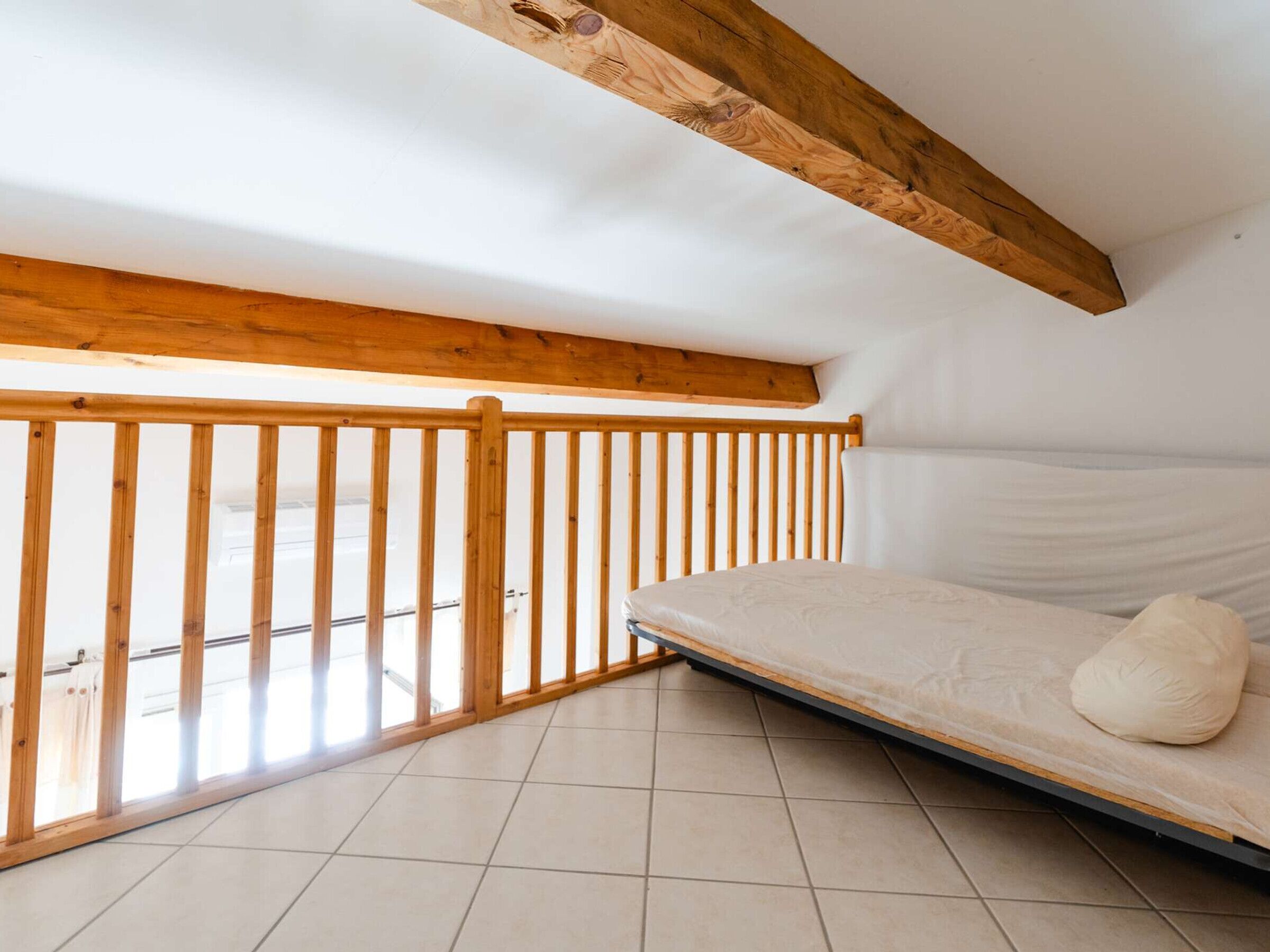 Een kleine slaapkamer op zolder, met schuine wanden, houten balken, een raam en een matras met kussens.