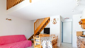 A cozy living room with a pink sofa, a wooden dining table with chairs, a staircase, a TV, and a kitchen area with cabinets.