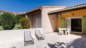 A sunlit patio with two lounge chairs, a table, and chairs, surrounded by patterned tiles and greenery.