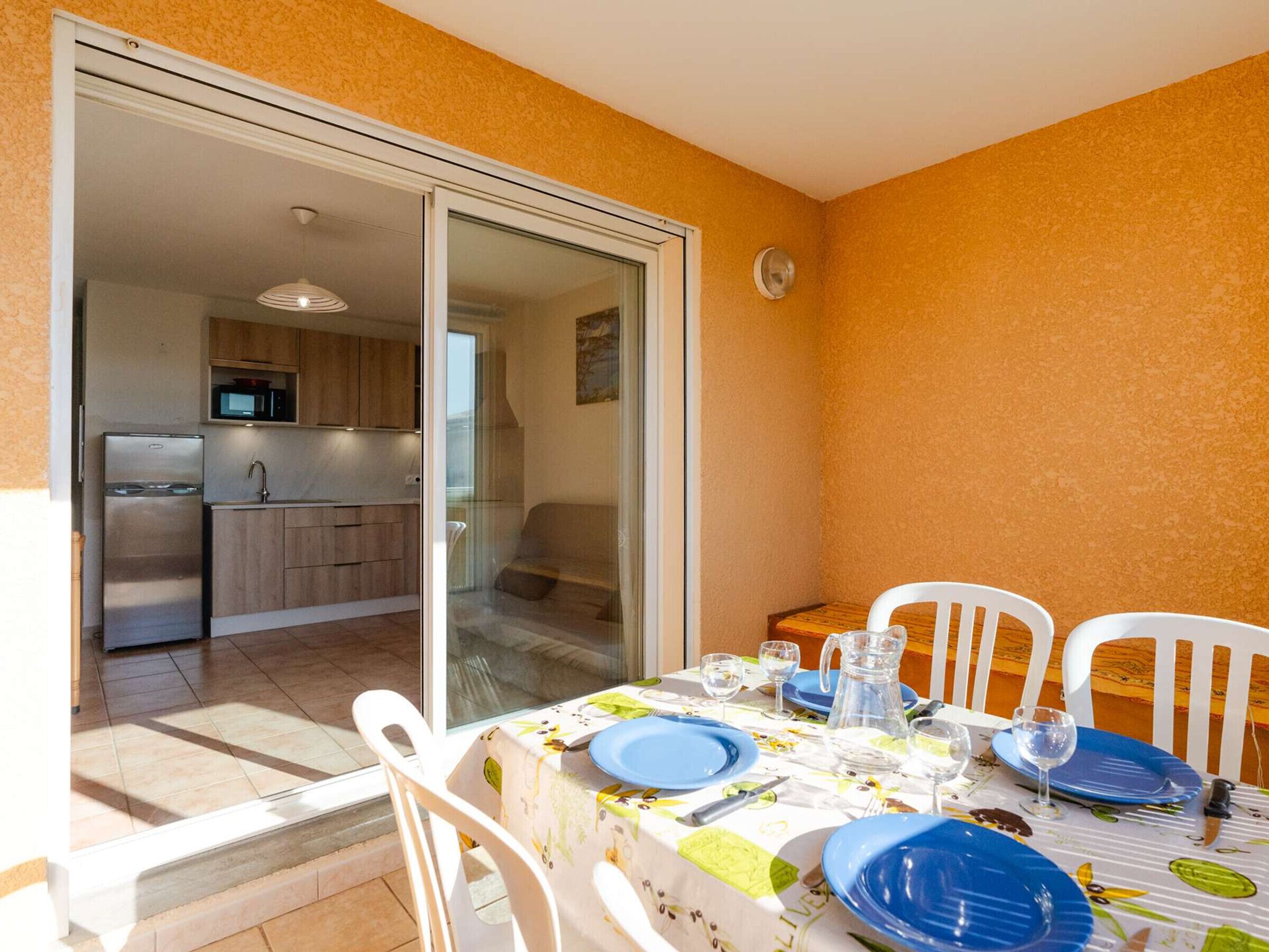 A dining area with a table set for four, a kitchen in the background, and a sliding glass door leading to an outdoor space.