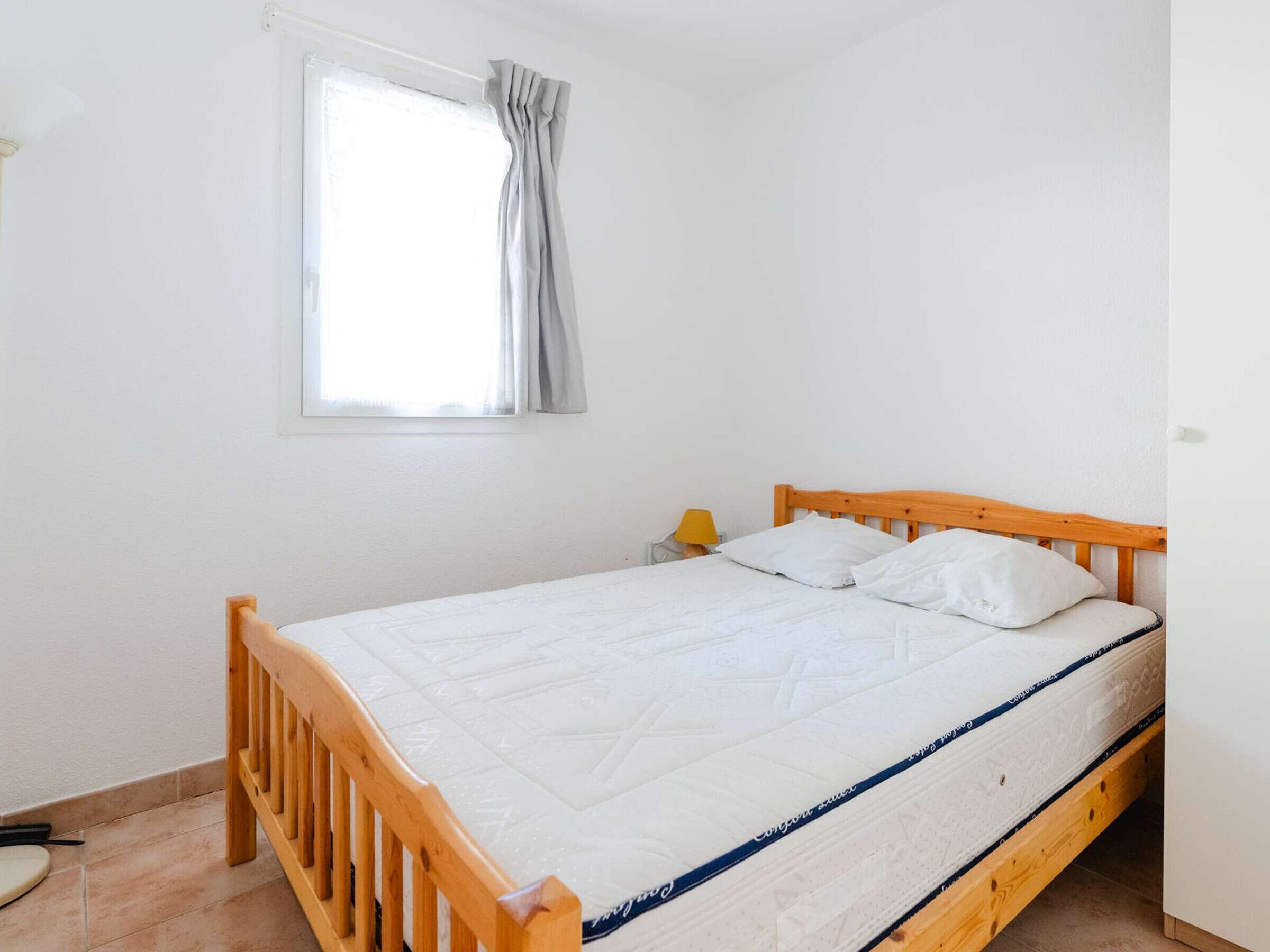 Un dormitorio con estructura de cama de madera, ropa de cama blanca, una mesita de noche con lámpara y una ventana con cortinas.