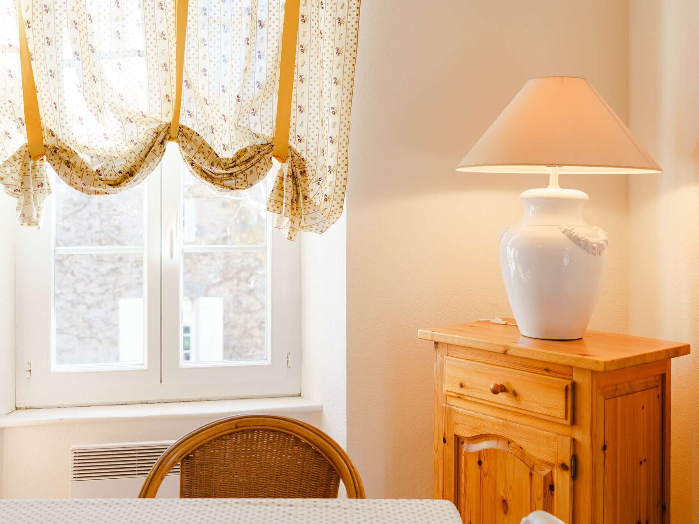 A room with a window, a lamp on a wooden cabinet, a wicker chair, and a table with a white tablecloth.