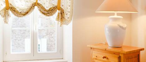 A room with a window, a lamp on a wooden cabinet, a wicker chair, and a table with a white tablecloth.