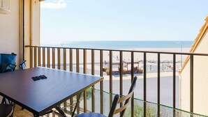 Un balcon avec une table et des chaises, donnant sur une plage et un parking.