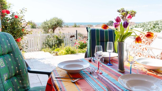 A dining table set outdoors with a view of the sea, a striped chair, and a vase with flowers.