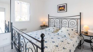Un dormitori amb un llit, un ventilador de sostre, una finestra amb cortines, una taula de nit amb una làmpada i un quadre amb marc a la paret.