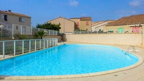 Una piscina amb un solàrium d'rajola, envoltada per una tanca, a prop d'edificis residencials amb persianes vermelles.