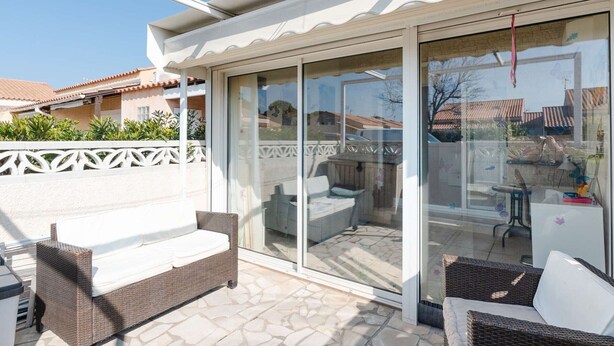 Ein überdachter Patio-Bereich mit Rattanmöbeln, eine Glastür zu einem Außenbereich und Blick auf benachbarte Häuser.