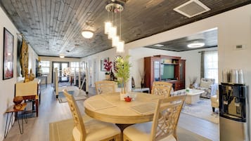 Ampla sala de estar com mesa de jantar redonda de madeira, várias cadeiras e uma cozinha moderna ao fundo.
