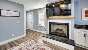 A modern living room with a fireplace, a mounted TV, and a kitchen area with white cabinets. - Peaceful Retreat Near Lake Barkley! (Cadiz)