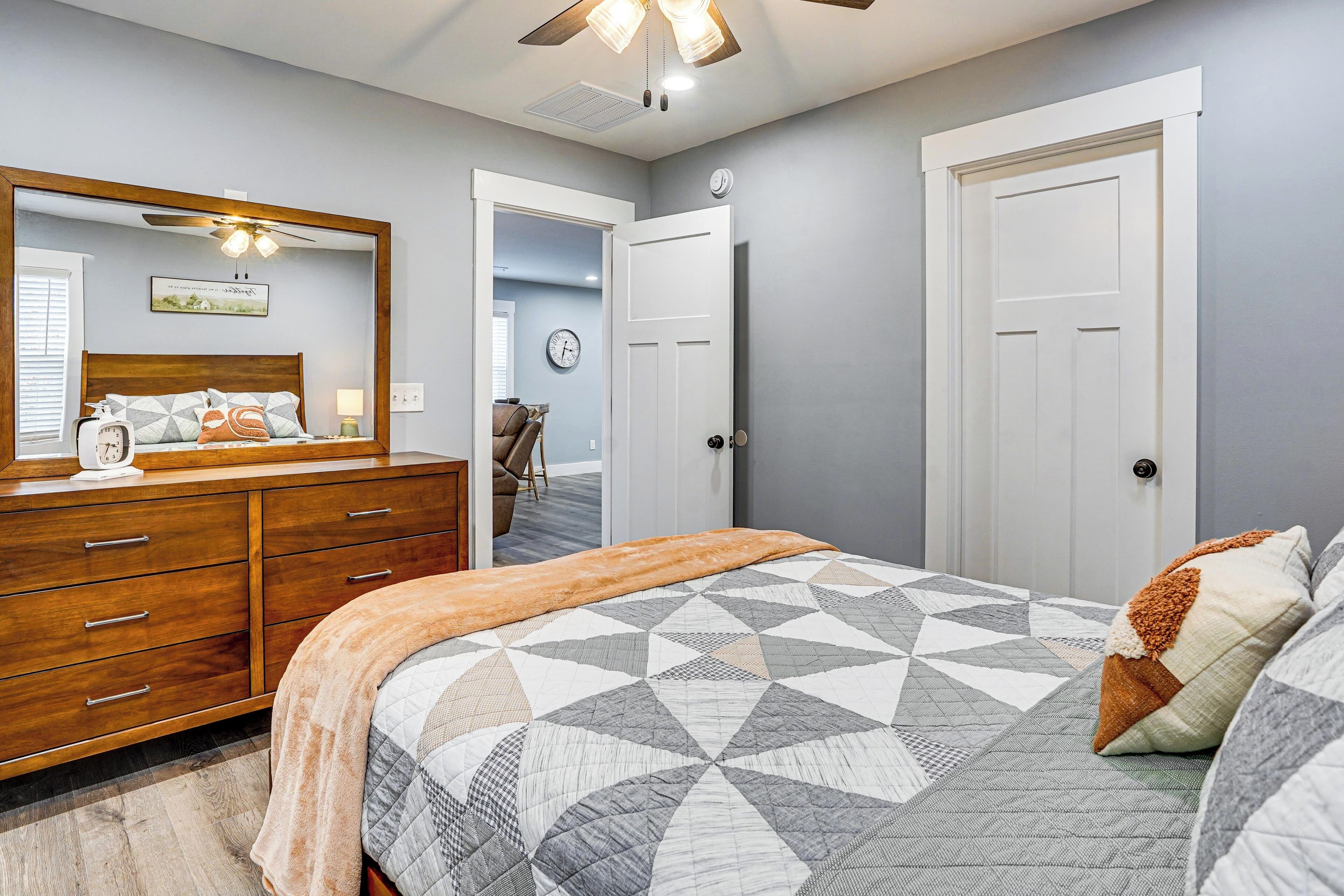 A bedroom with a wooden dresser, a bed with a geometric patterned quilt, a ceiling fan, and a door leading to another room.