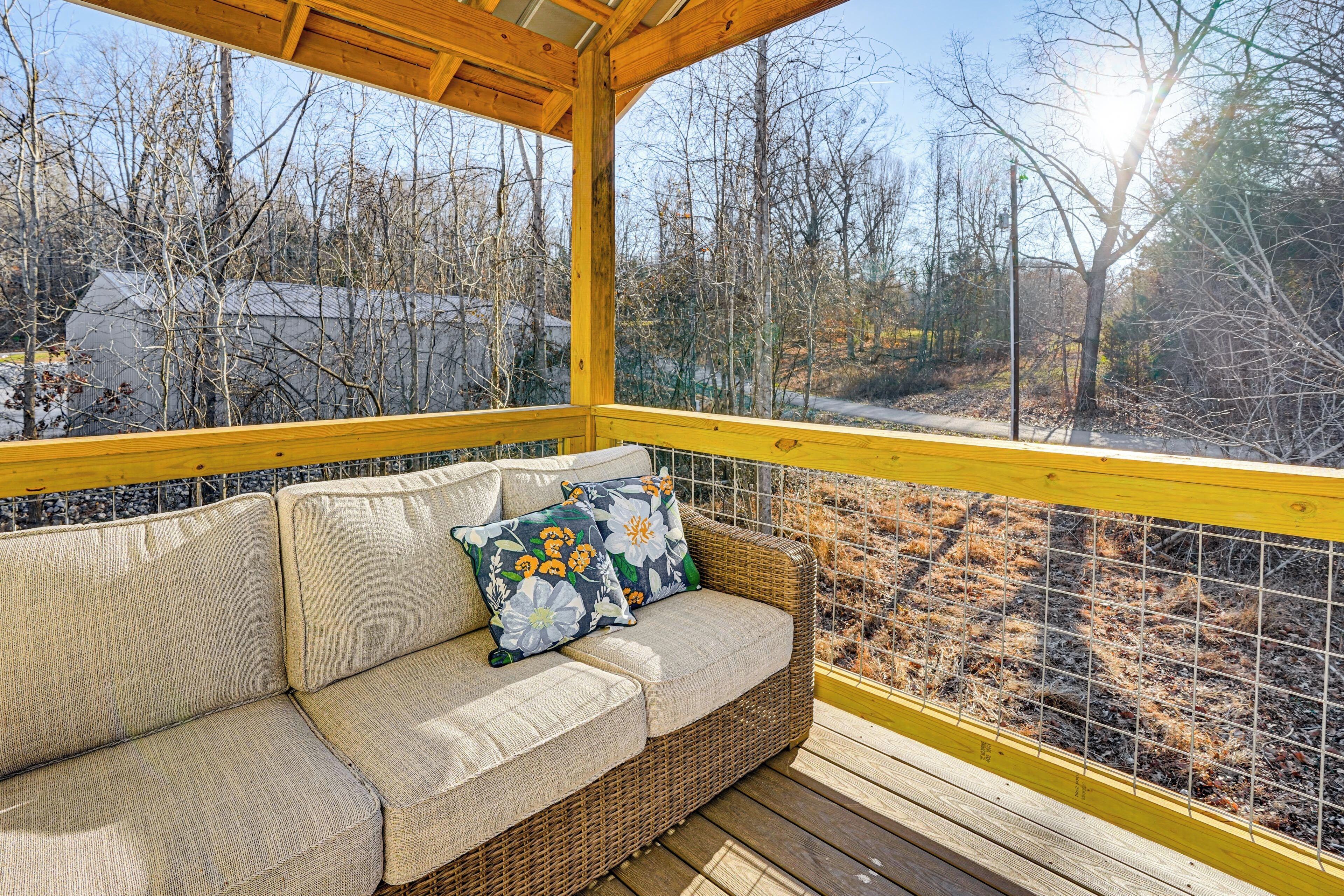 A covered outdoor seating area with a wicker sofa and cushions, a yellow railing, and a view of trees and buildings.