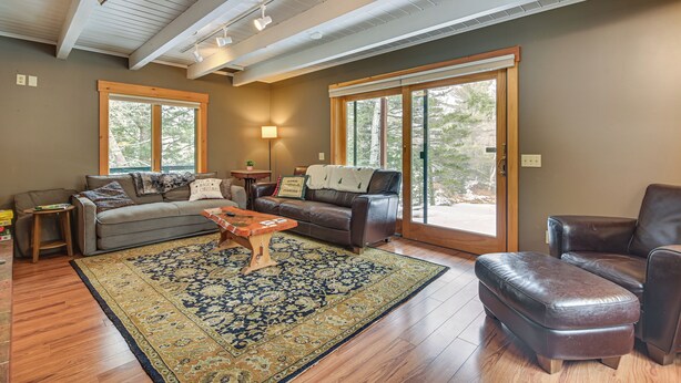 A living room with a sofa, ottoman, coffee table, and a large rug. There are two windows and a door leading outside. - Riverfront! All-season Base 11 Mi to Sugarloaf Mtn (Kingfield)