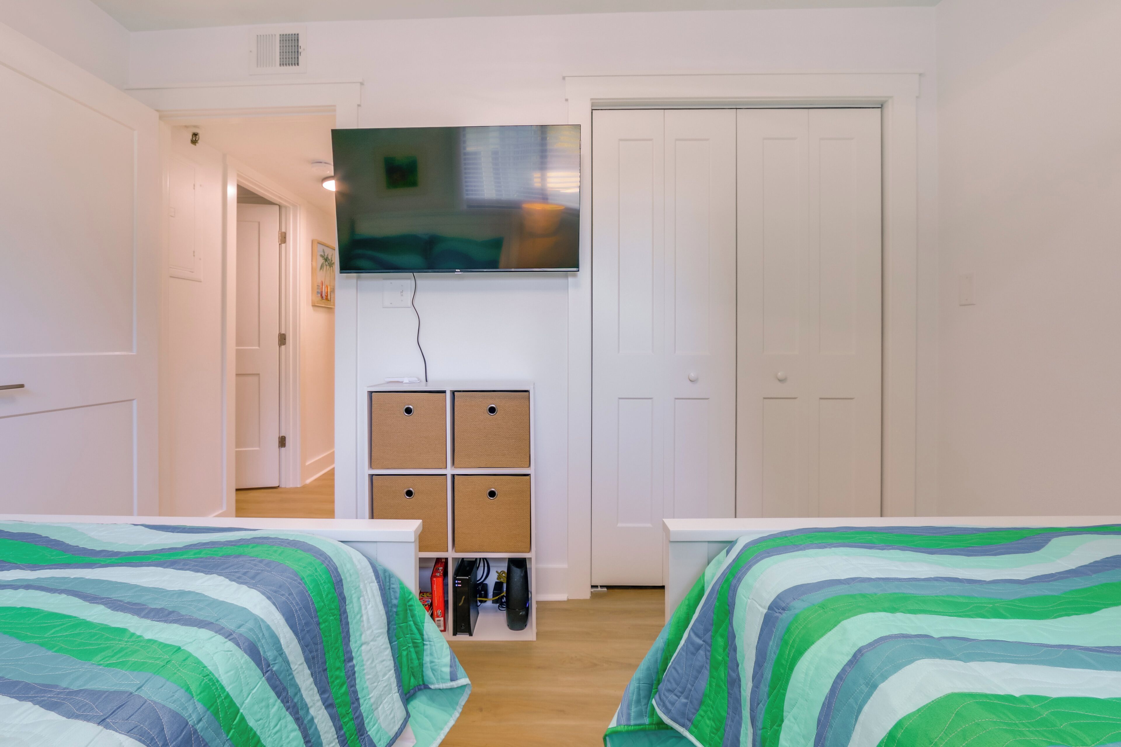 A hotel room with two beds, a TV mounted on the wall, a storage unit below the TV, and a door leading to another room.