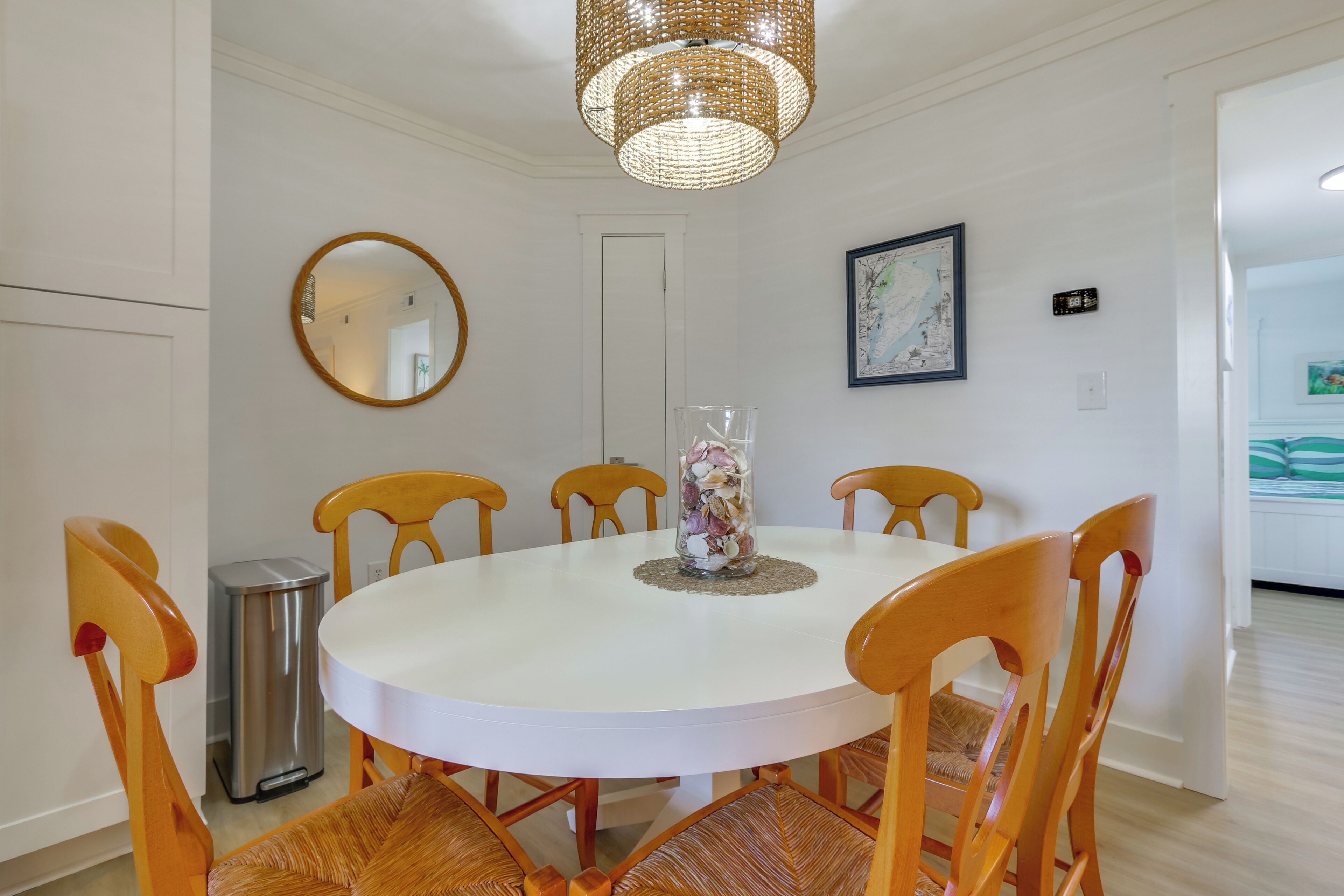 Un coin repas comprenant une table ronde blanche, six chaises en bois, une poubelle, un miroir et un luminaire suspendu.