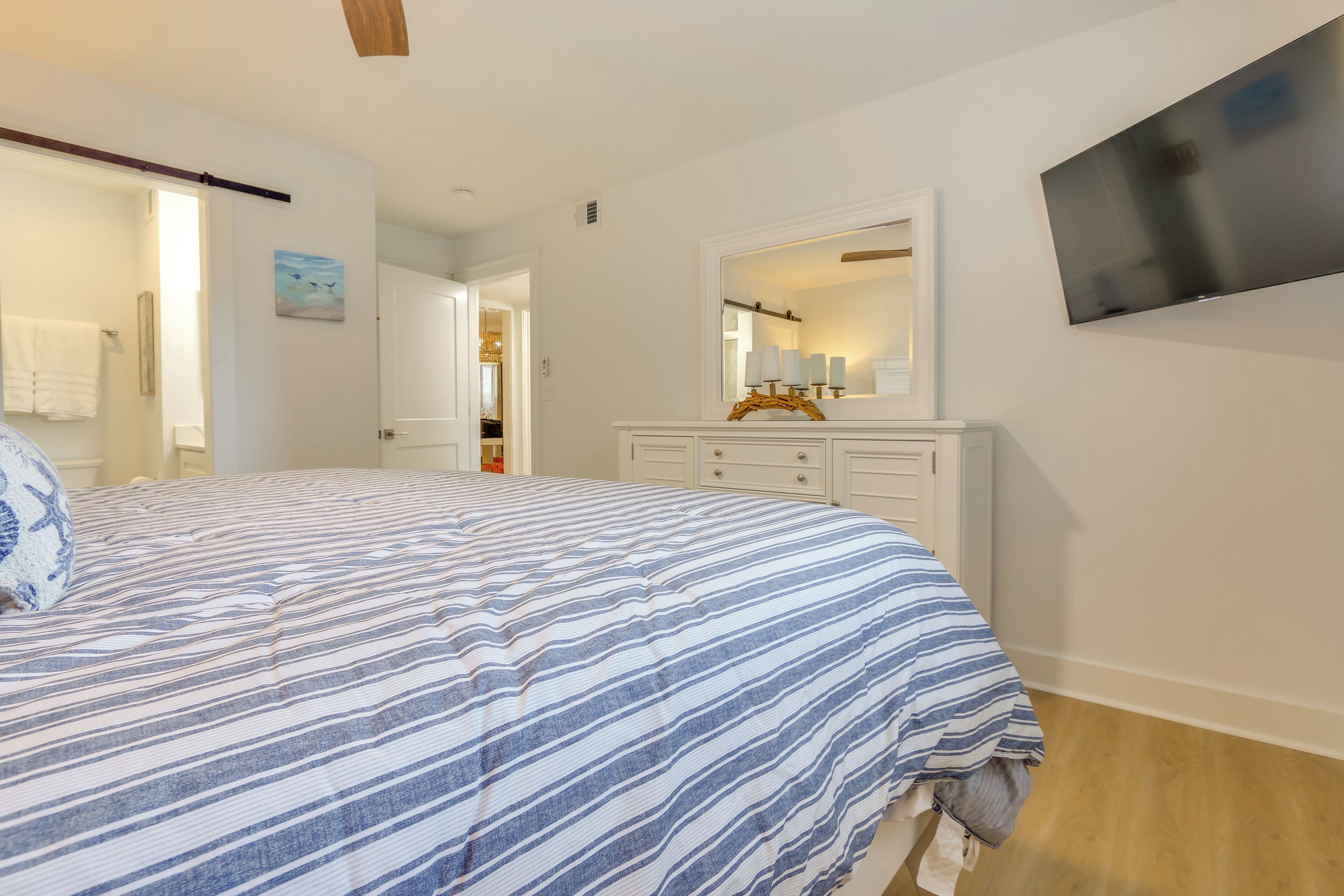 A bedroom with a large bed, a dresser, a TV mounted on the wall, and a door leading to another room.