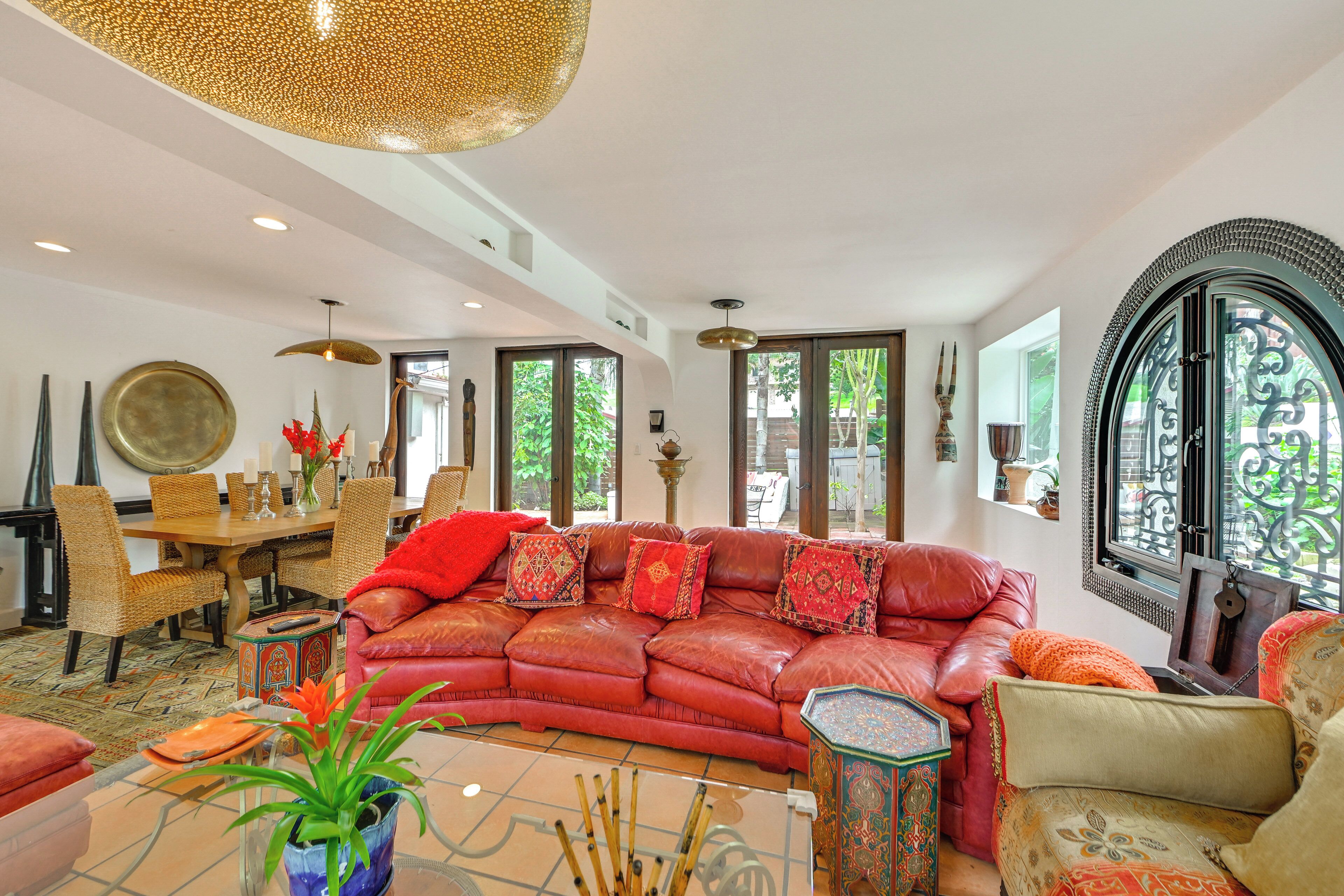 A spacious living room with a red leather sofa, a dining area with wicker chairs, and a large window with a view of greenery.