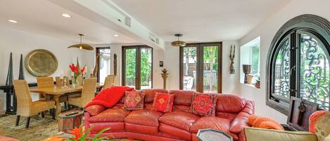 A spacious living room with a red leather sofa, a dining area with wicker chairs, and a large window with a view of greenery.