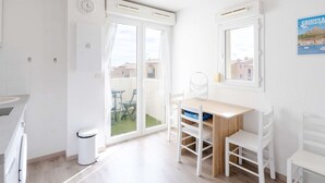 Una habitación luminosa y blanca con una mesa y sillas de madera, una ventana con vistas a edificios y un reloj en la pared.