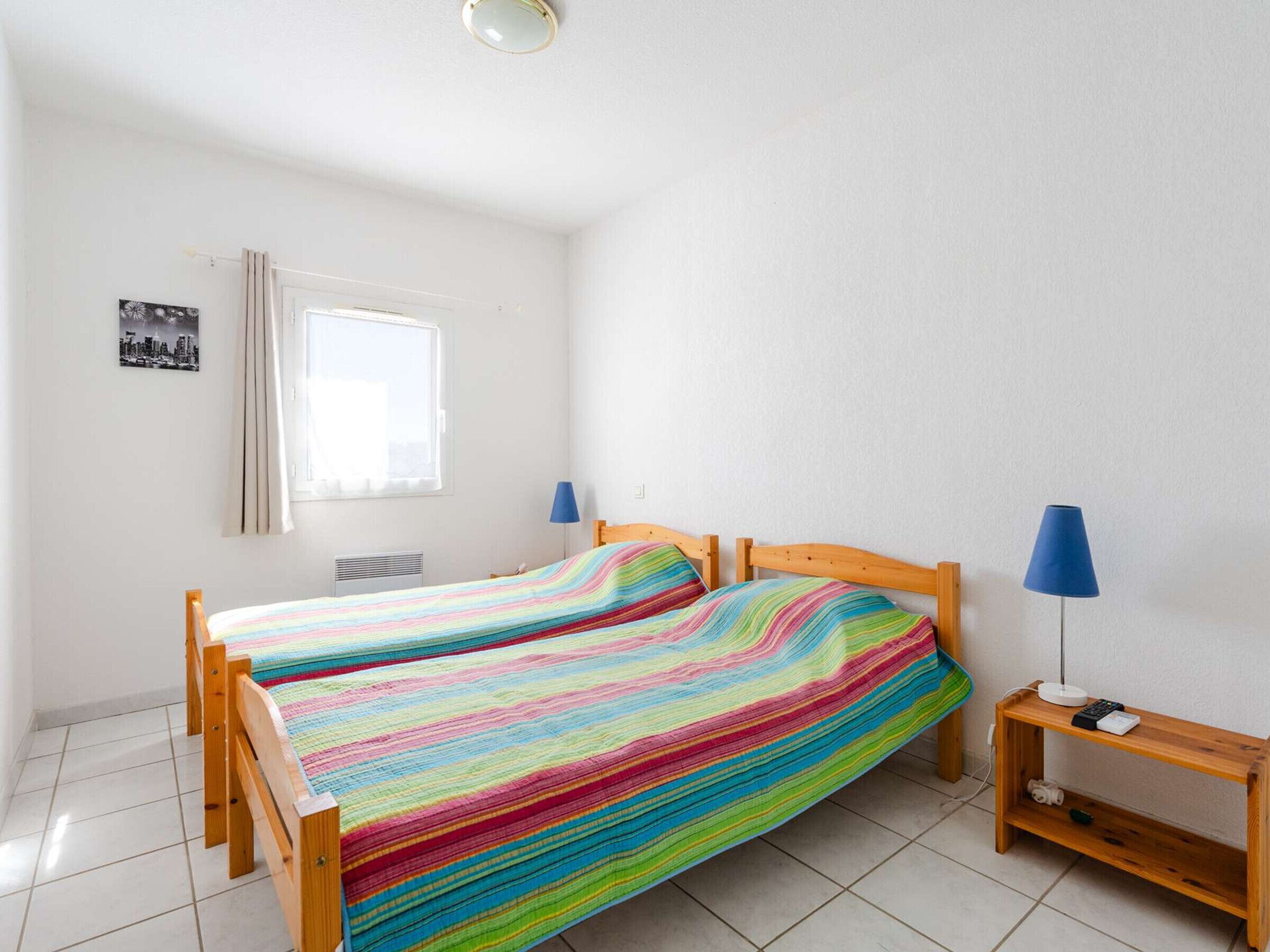 Ett rum med två sängar i trä, ett fönster med gardiner, en tavla som hänger på väggen och ett nattduksbord med en lampa.