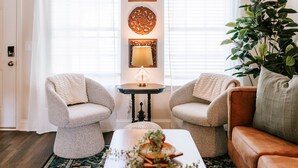 Un salon chaleureux avec deux fauteuils, une table basse et un canapé. On y trouve une lampe sur une petite table, une grande plante et des œuvres d’art décoratives accrochées au mur.