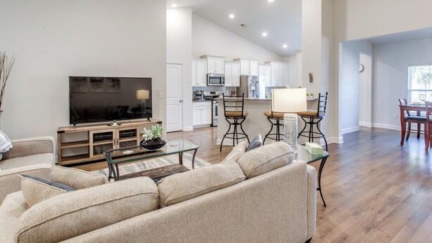 A spacious living room with a large sofa, a flat-screen TV, and a coffee table. There is a kitchen area with white cabinets and a dining area with a table and chairs. - Ultimate Golf Resort Getaway in Myrtle Beach! (Myrtle Beach)