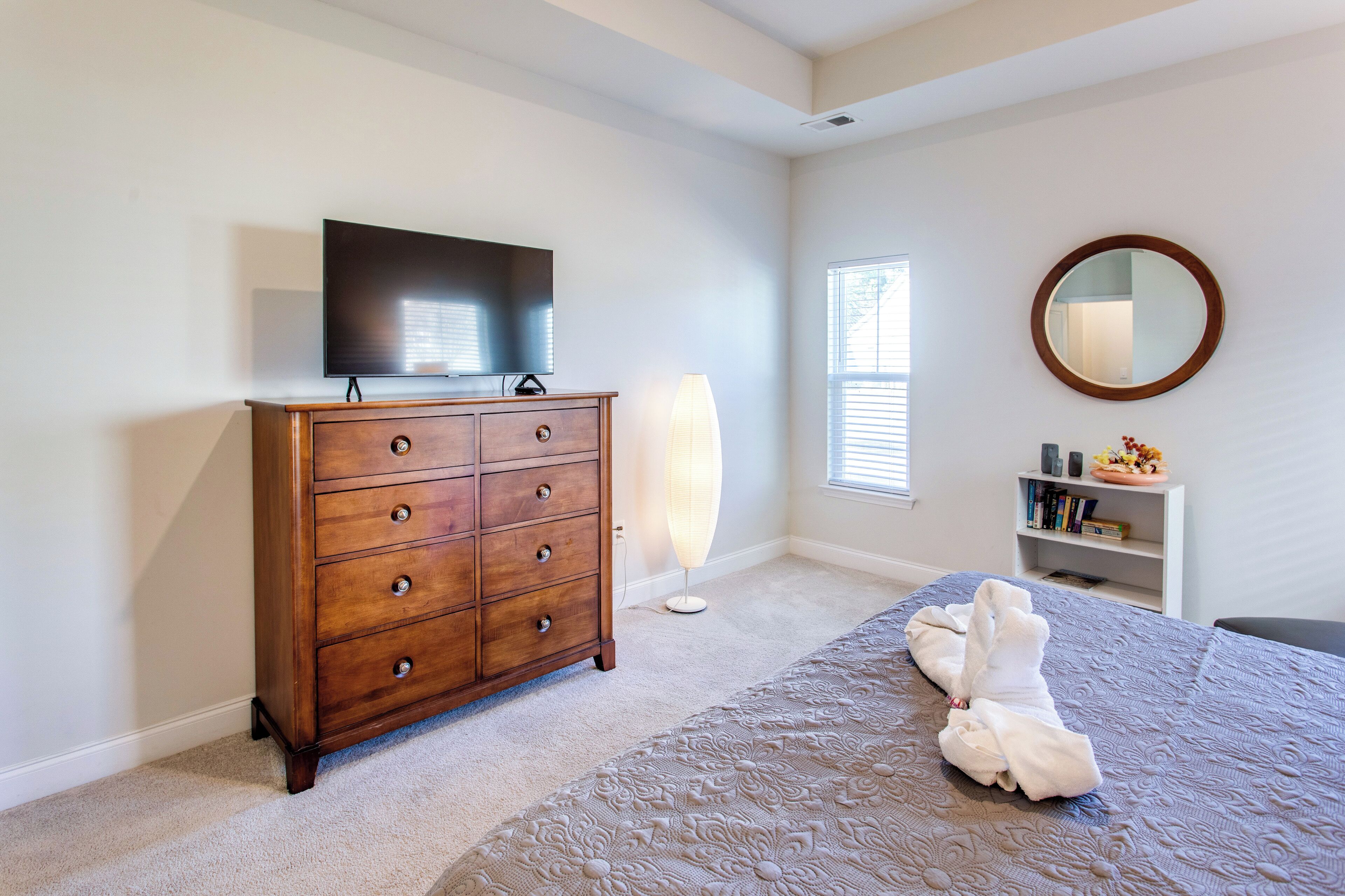 A bedroom with a wooden dresser, a flat-screen TV, a bedside lamp, a round mirror, and a neatly made bed.