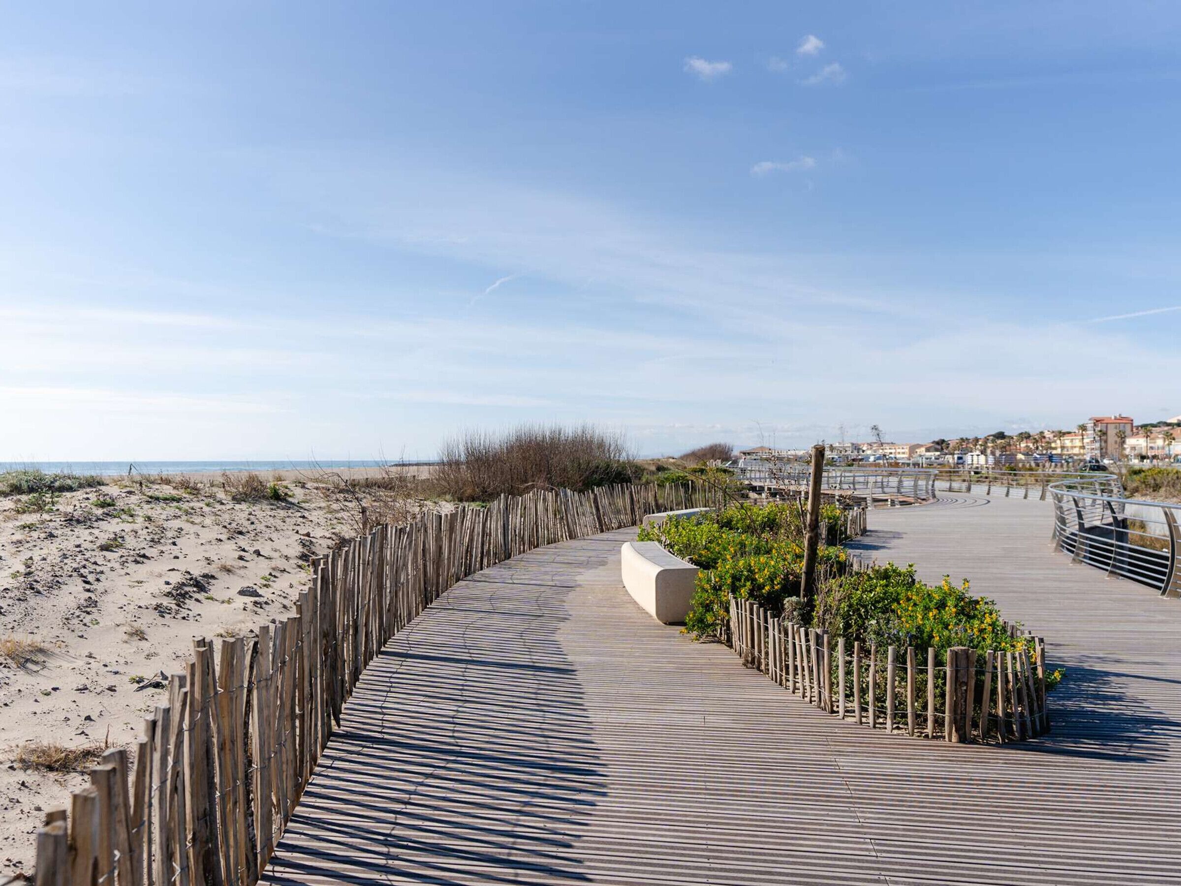 Een houten looppad langs een strand, met een gebogen ontwerp, dat uitkomt op een verhard gebied met leuningen en groen.