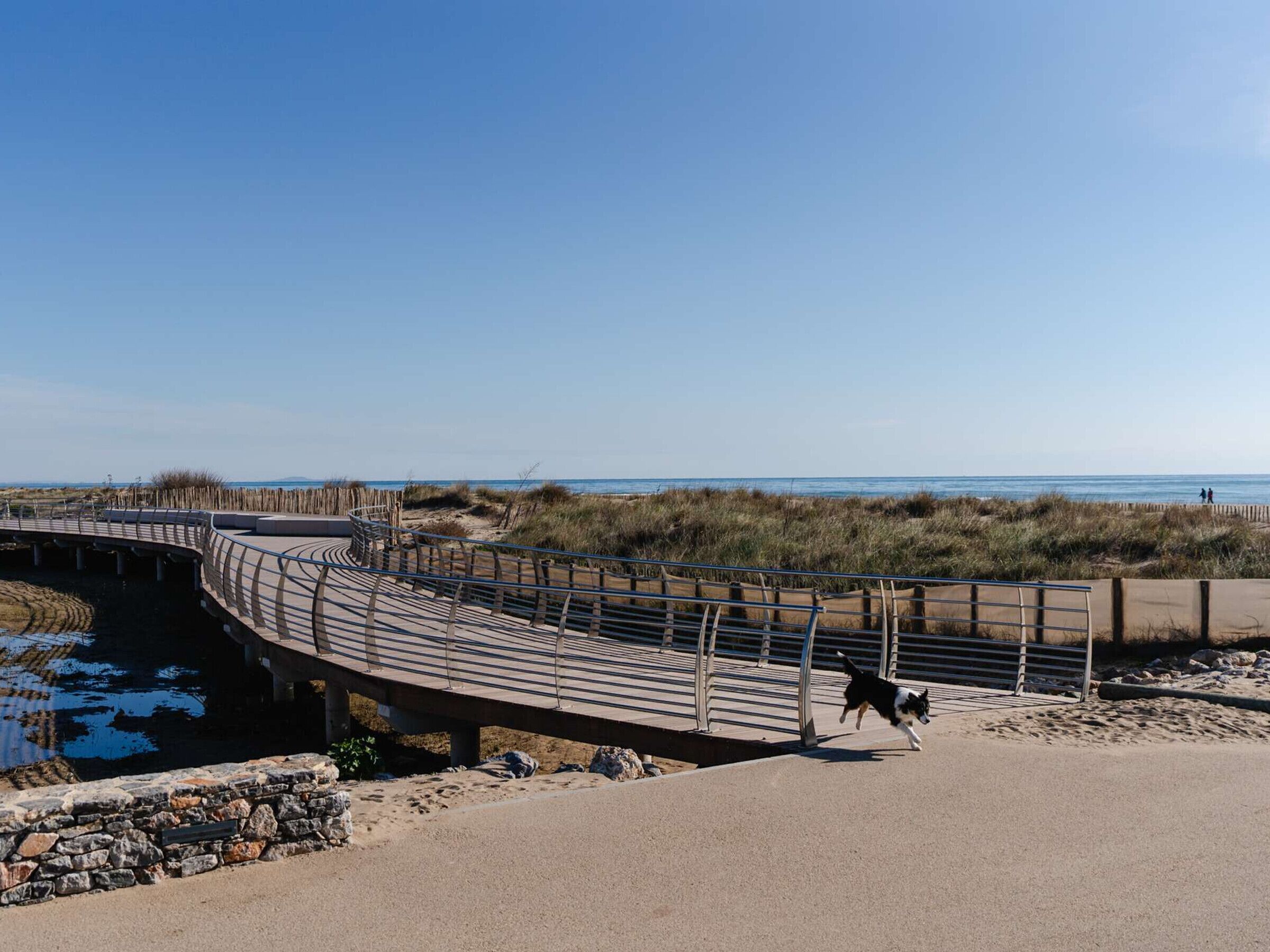Una passerella di legno curva che conduce a una spiaggia con dune e cielo sereno.