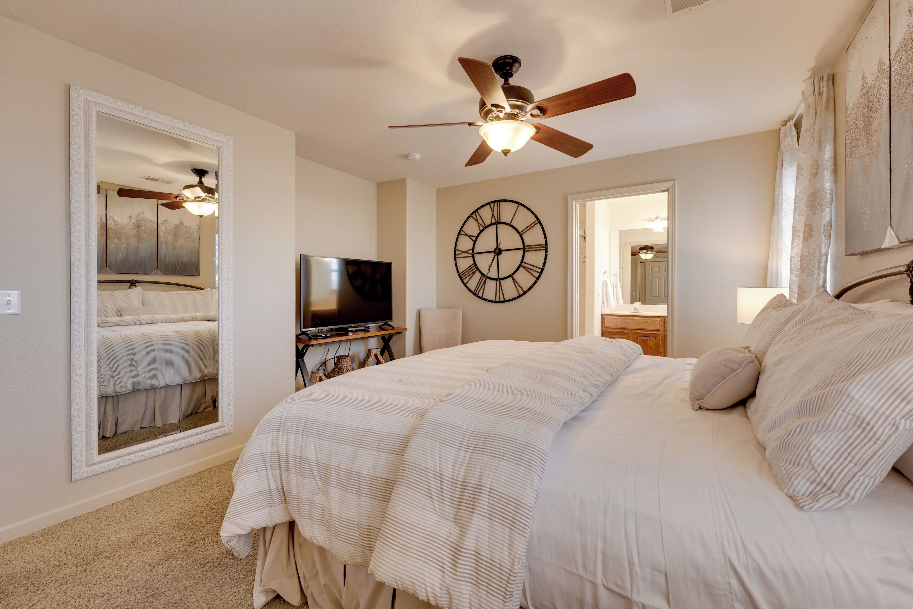 A bedroom with a large bed, a ceiling fan, a wall clock, a television, and a mirror.