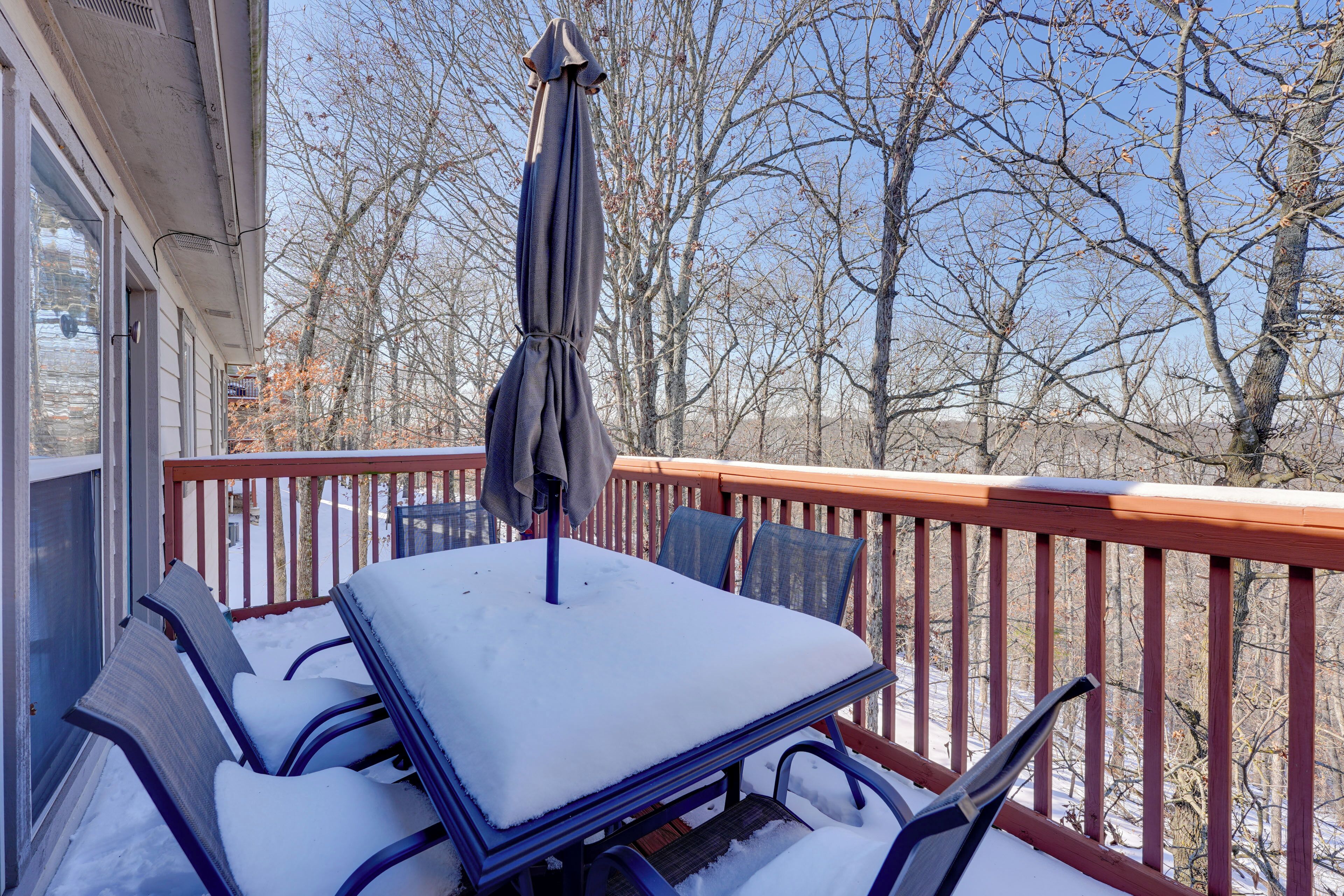 A snow-covered deck with a table and chairs, a wooden railing, and leafless trees in the background.