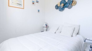 A bedroom with a white bed, a nightstand with a lamp, and a wall with framed pictures and a decorative blue and gold wall art piece. - 1-bedroom apartment with balcony, private parking, pets allowed (Fleury-Saint-Pierre-la-Mer)