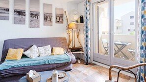 A cozy living room with a sofa, a table with a tissue box and a vase, a lamp, and a view of a balcony with a table and chairs. - 1-bedroom apartment with balcony, private parking, pets allowed (Fleury-Saint-Pierre-la-Mer)