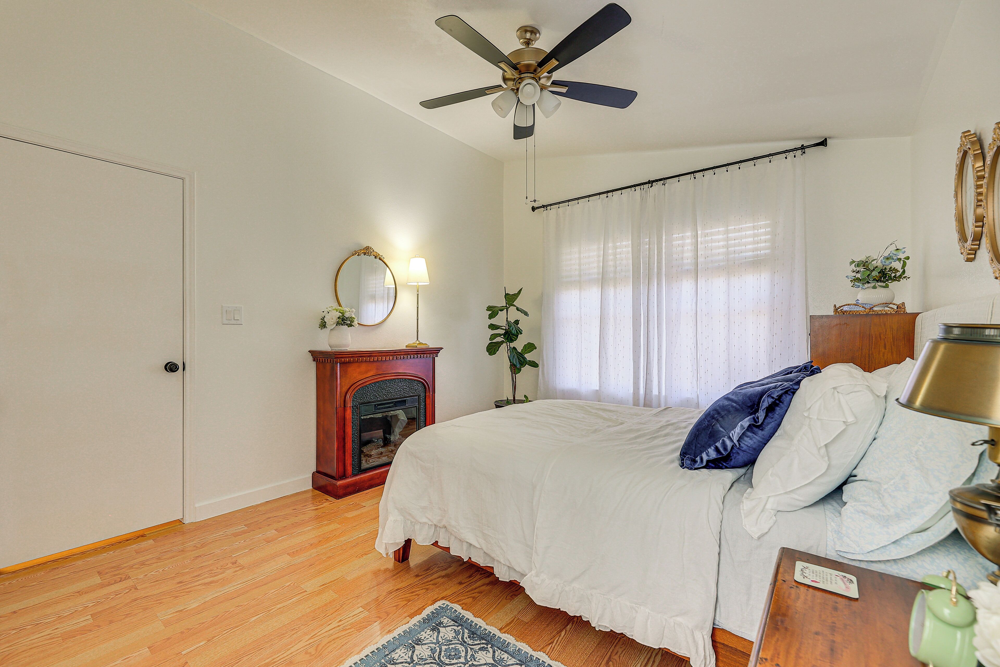 A bedroom with a bed, a ceiling fan, a fireplace, a nightstand, and a window with curtains.