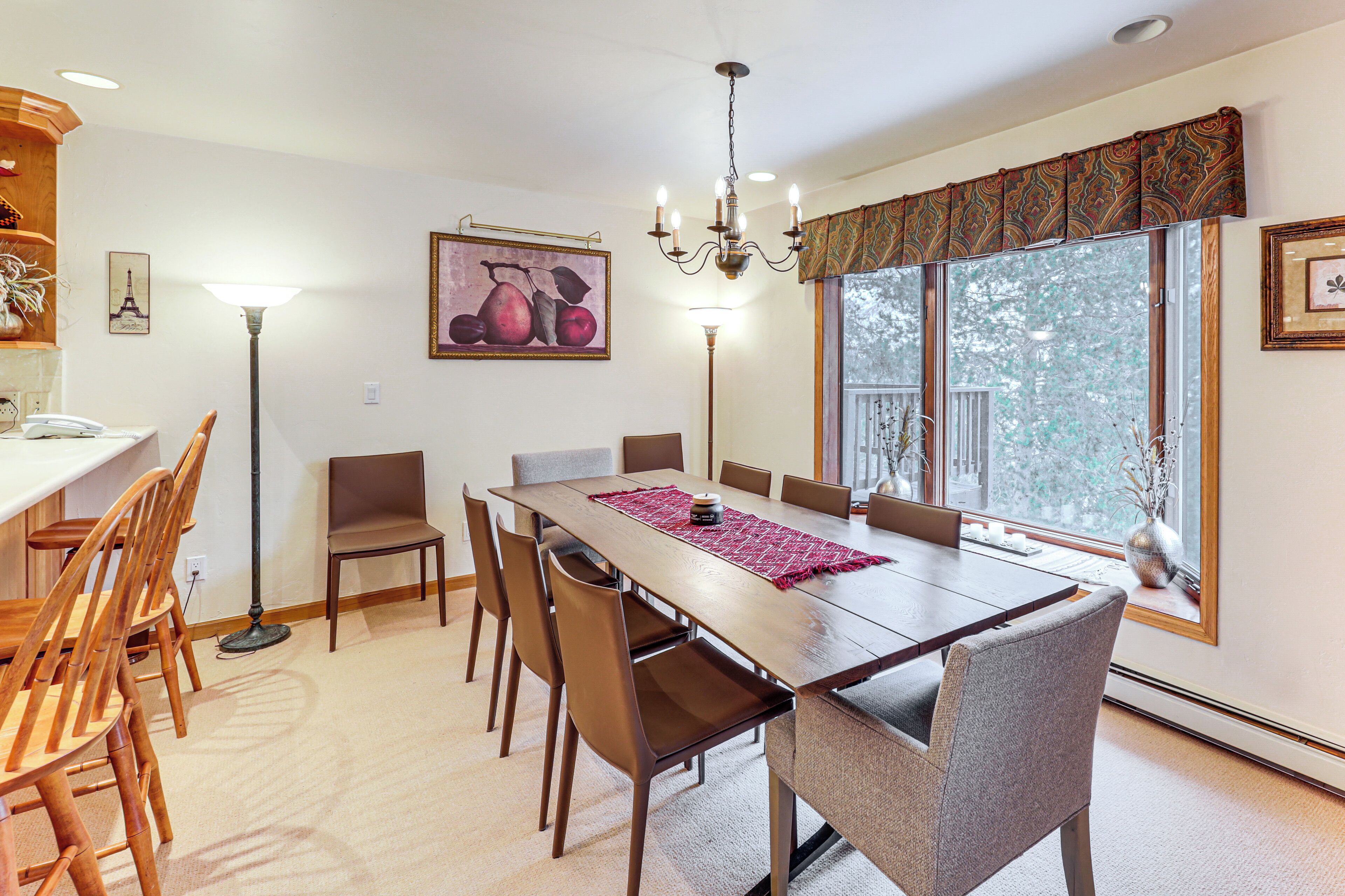 A dining area with a long table, multiple chairs, a chandelier, and a large window with a view of trees.