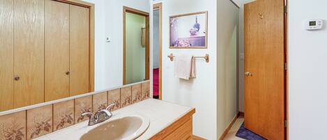 Un baño con tocador de madera, lavamanos, espejo y puerta que da a otra habitación.