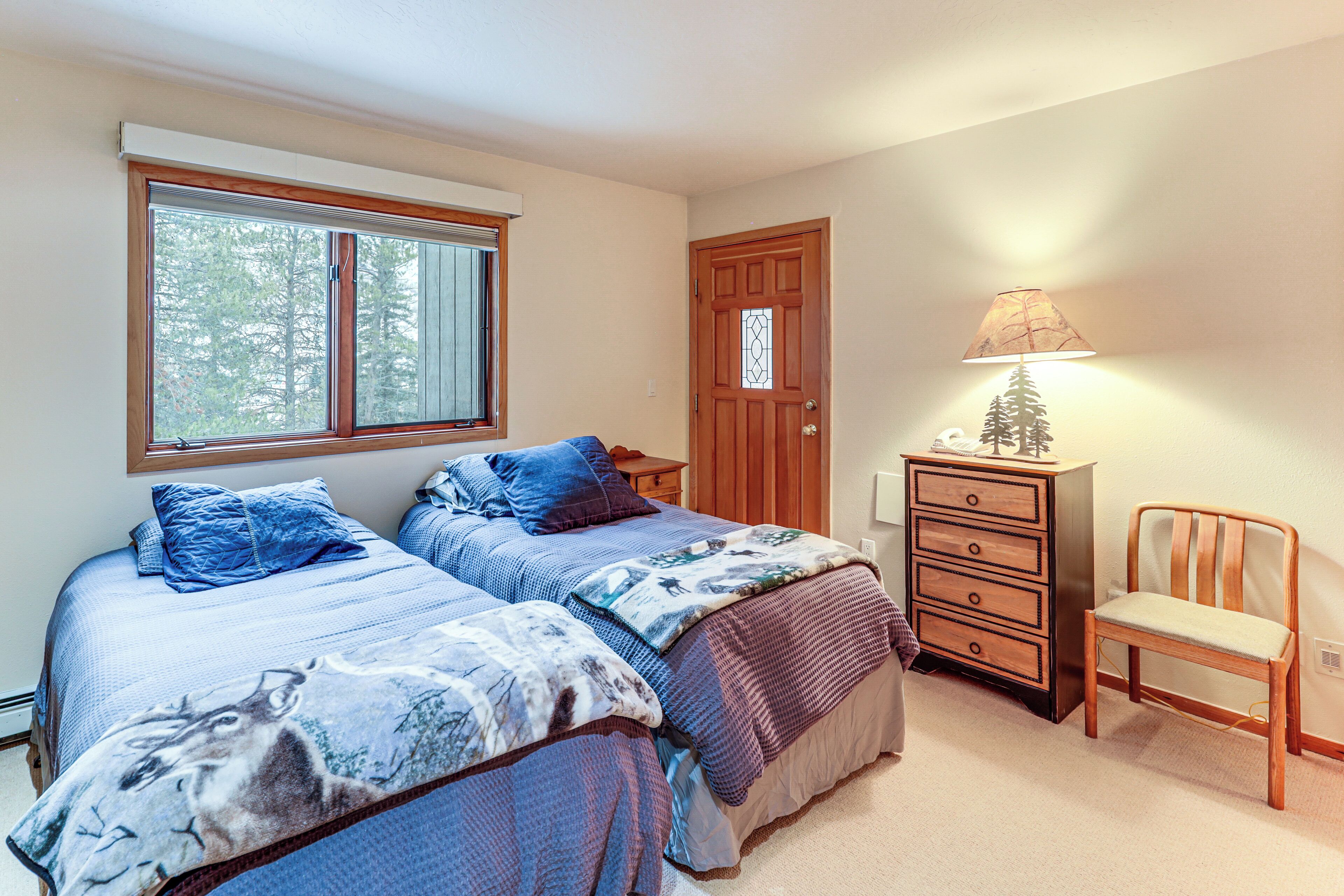 A bedroom with two beds, a dresser, a chair, a window with blinds, and a door with a glass panel.