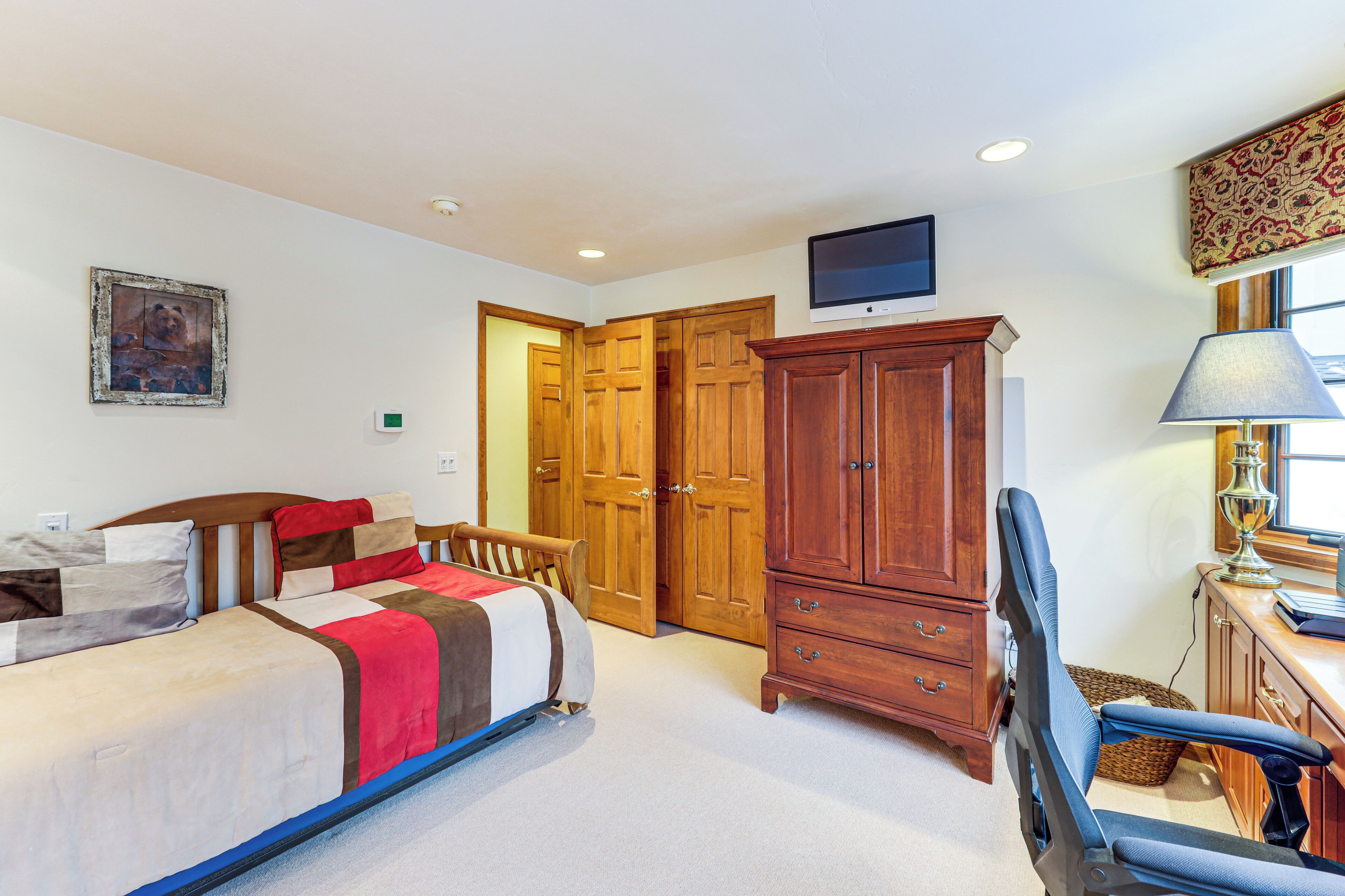 A bedroom with a bed, wooden wardrobe, desk, chair, and a television on the wall.
