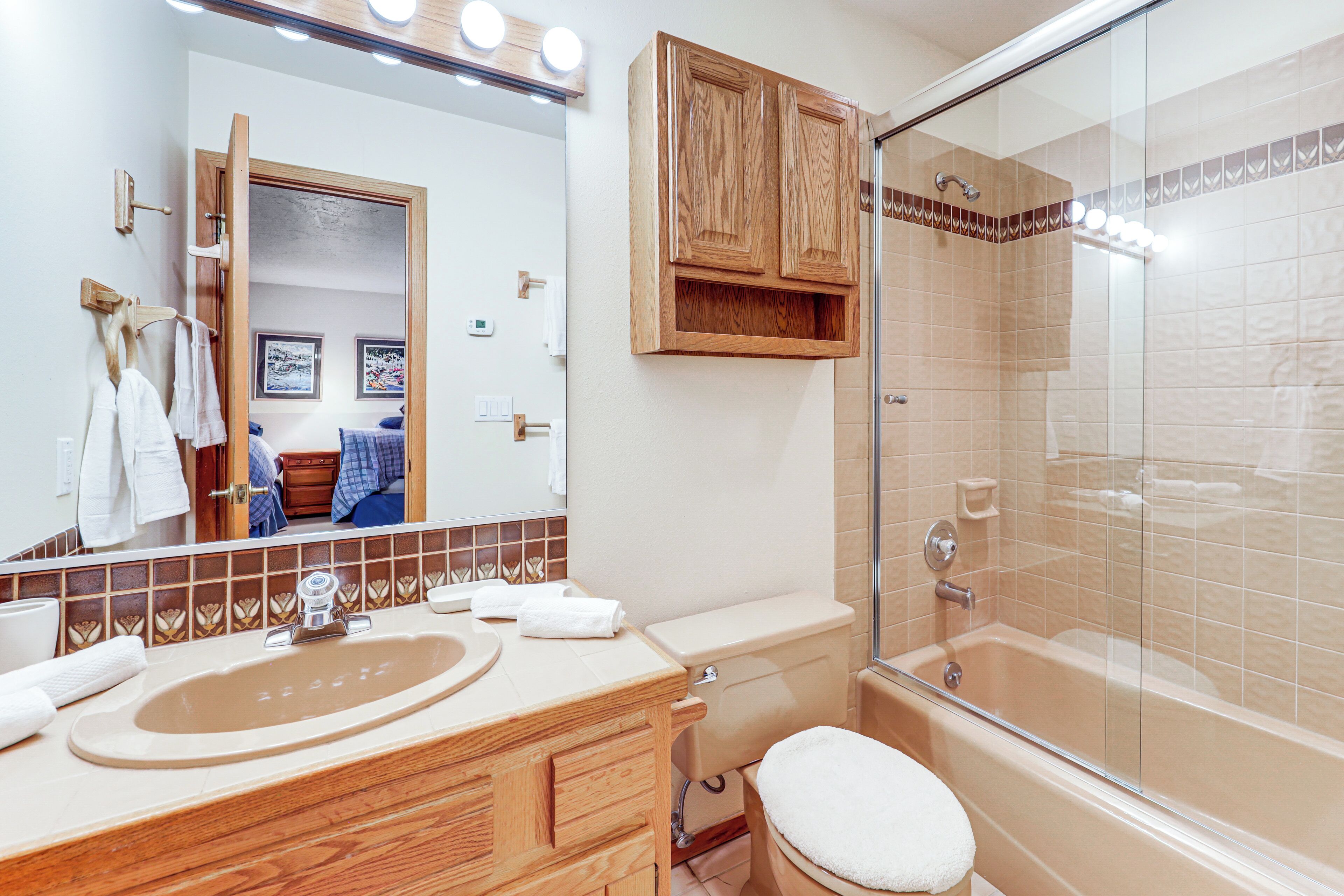 Un baño con tocador de madera, inodoro, bañera con mampara de vidrio y espejo con luces superiores.