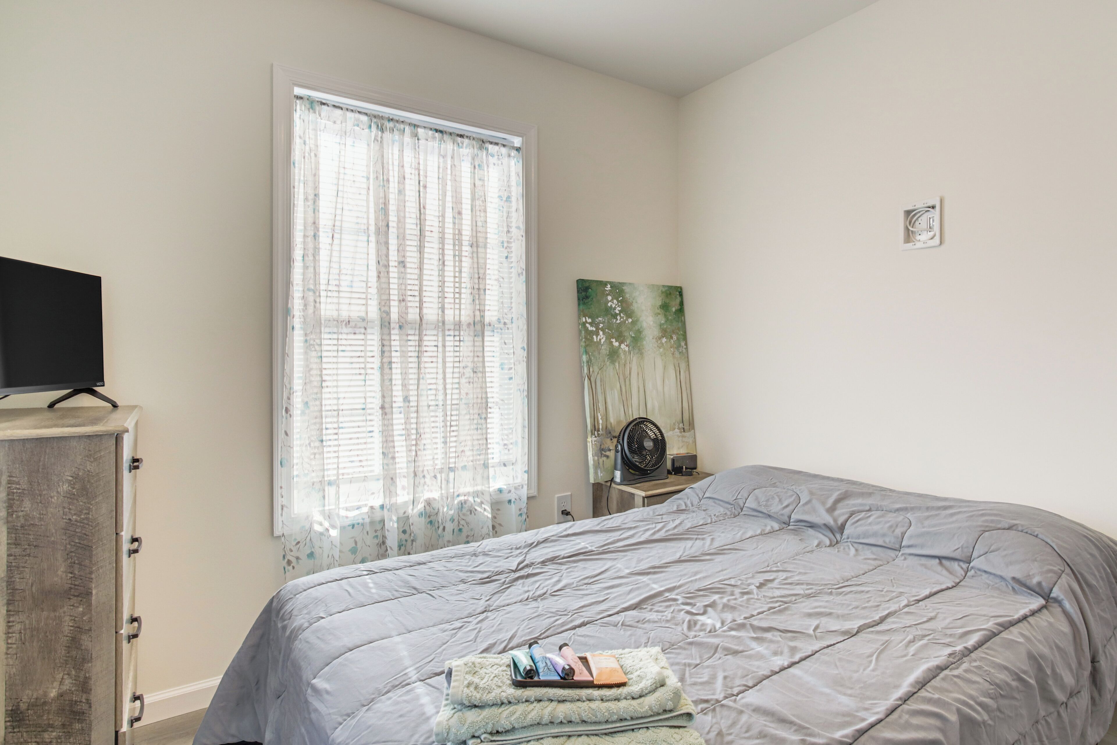 A bedroom with a bed, a dresser, a window with curtains, and a wall-mounted fan.