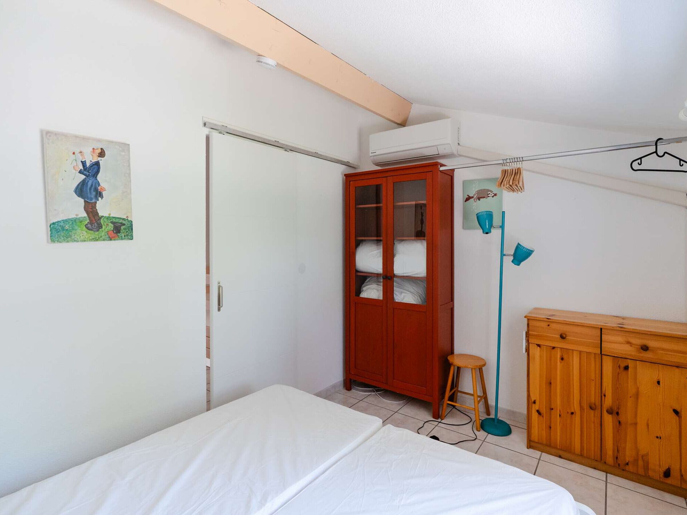 Un dormitorio con cama, armario, taburete, lámpara colgante y estante en la pared.