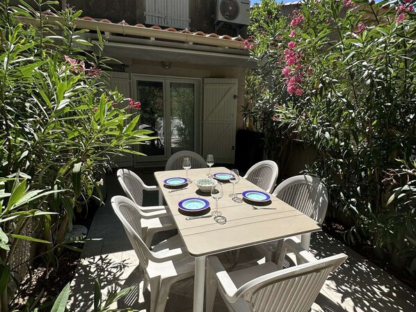 Une terrasse extérieure avec une table dressée pour quatre personnes, entourée de chaises blanches et d’une végétation luxuriante.