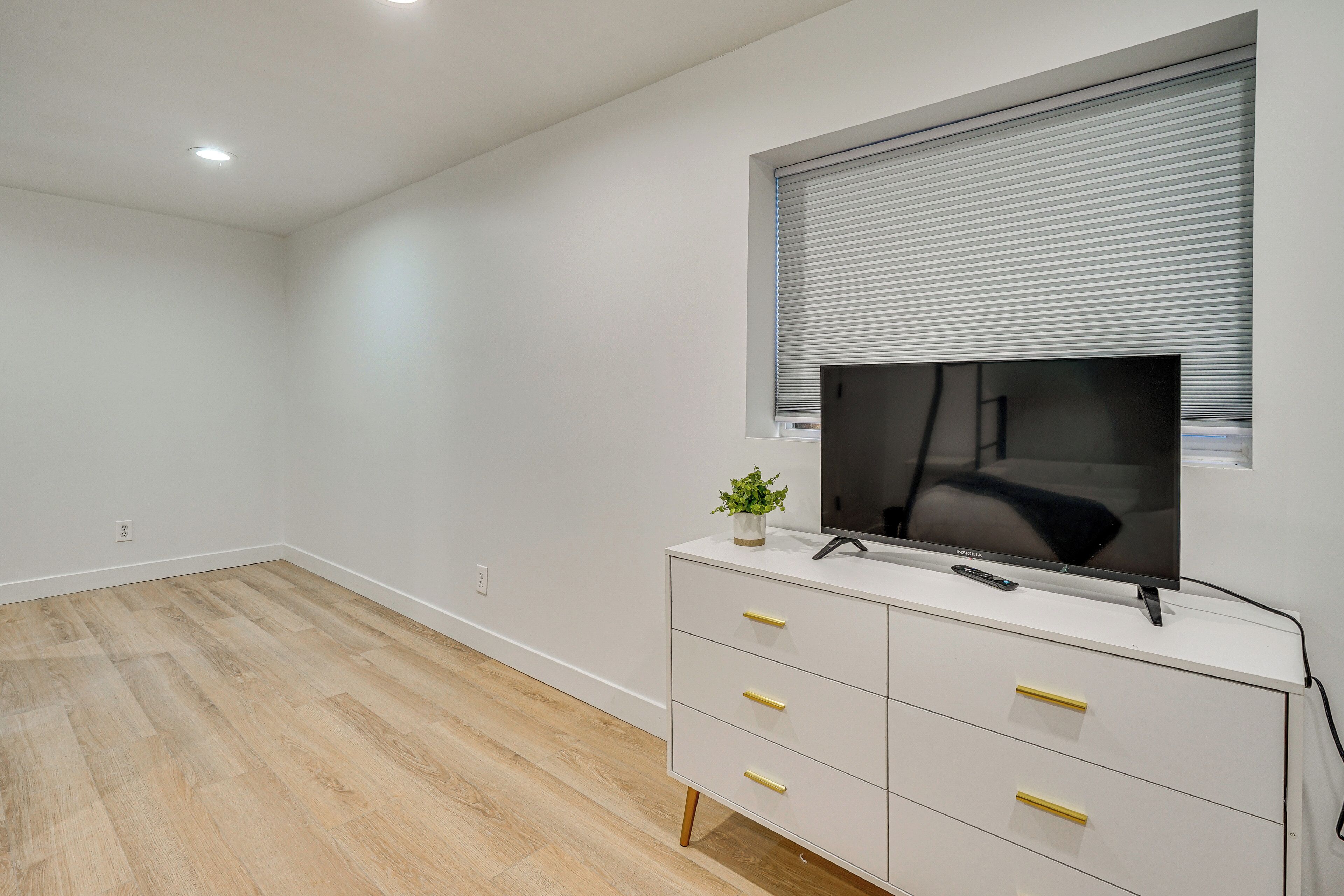 Una sala moderna con un televisor de pantalla plana montado en la pared, un mueble blanco con tiradores dorados y una ventana con persianas.