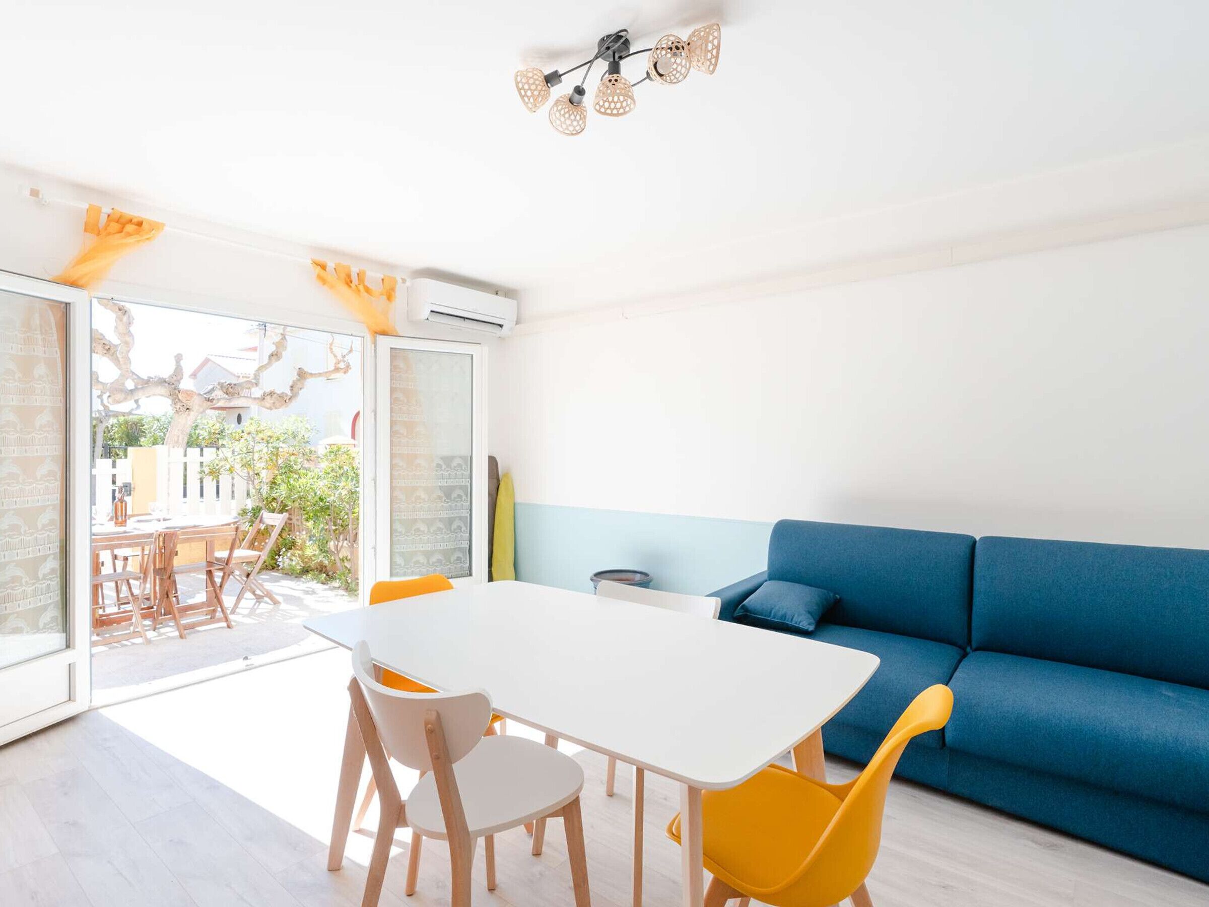 Un salon moderne avec une table blanche, des chaises jaunes, un canapé bleu et une porte en verre donnant sur un espace extérieur.