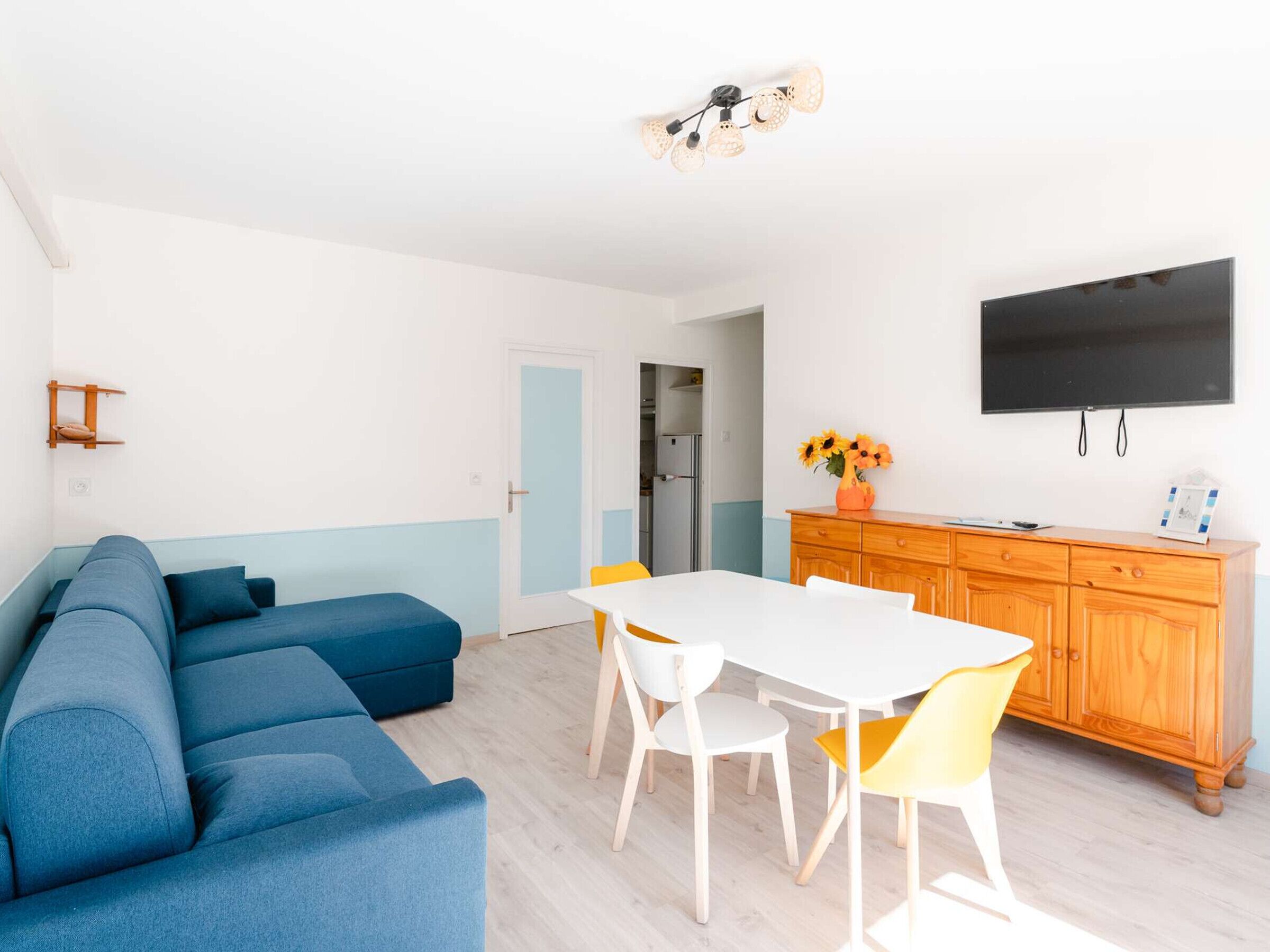 Ein modernes Wohnzimmer mit einem blauen Ecksofa, einem weißen Esstisch mit gelben Stühlen, einer Holzkommode und einem an der Wand befestigten Flachbildfernseher.