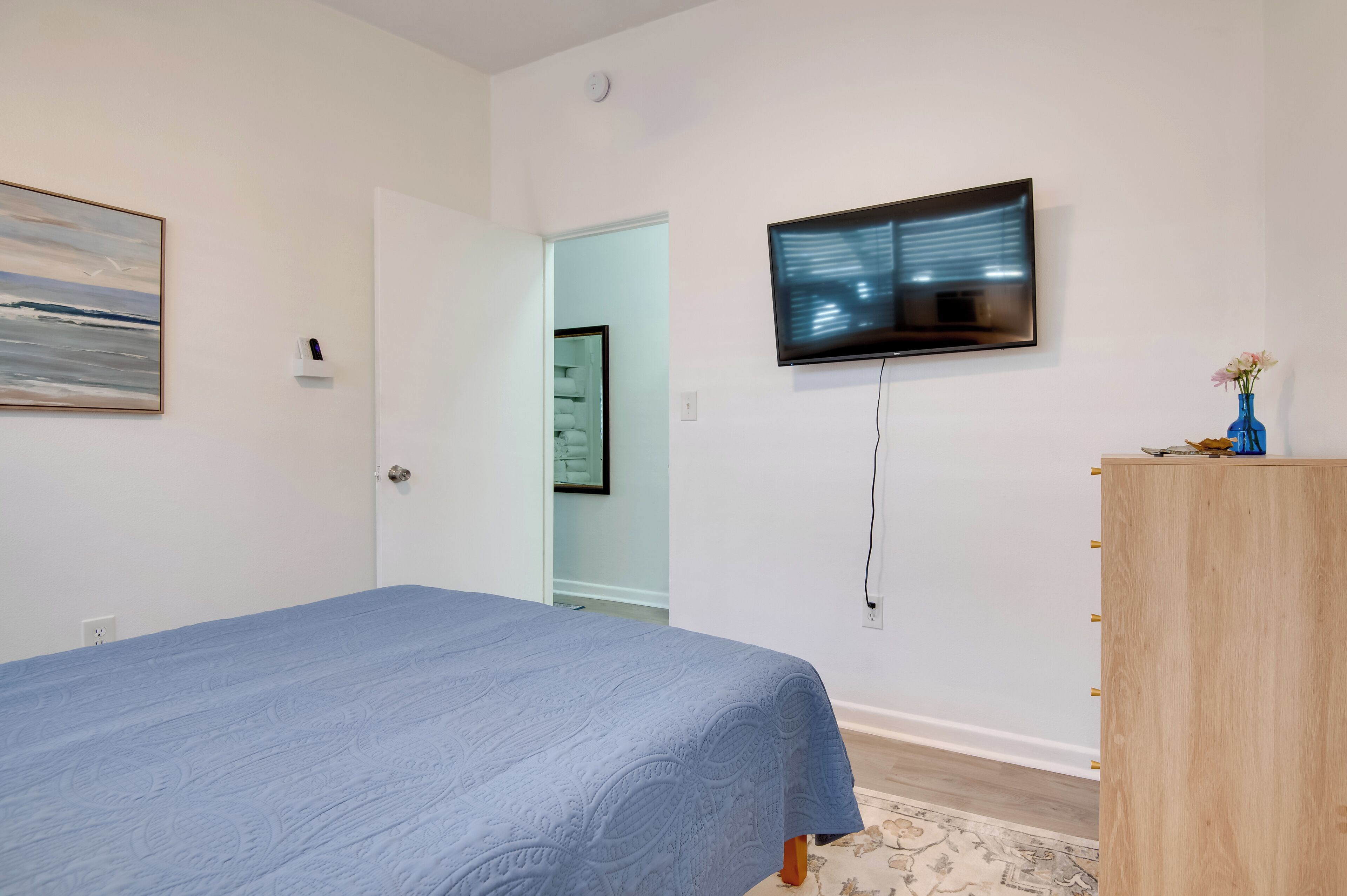 A bedroom with a bed, a TV mounted on the wall, a wooden dresser, and a door leading to another room.