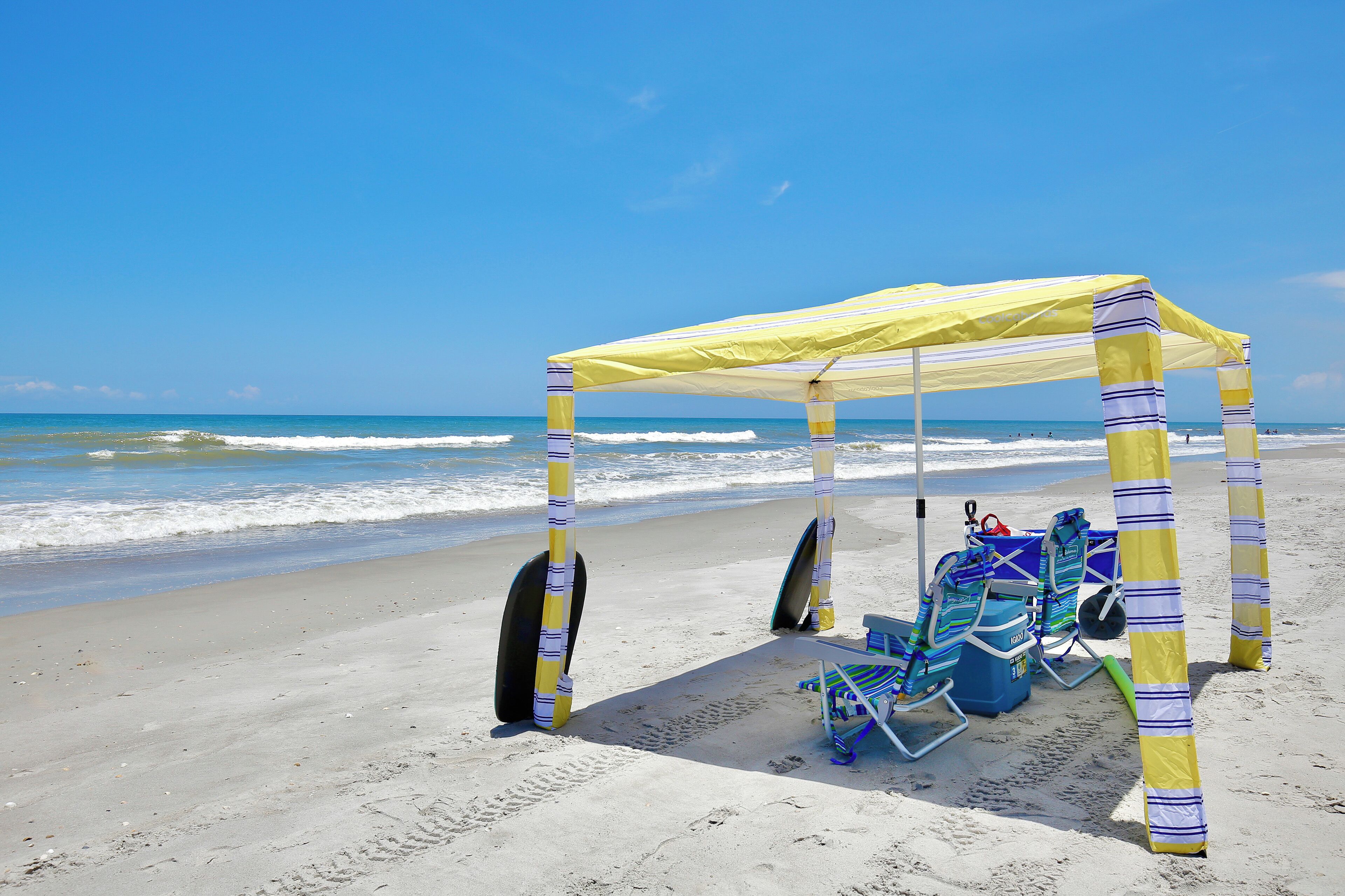 Une plage avec un parasol à rayures jaunes et blanches, des chaises de plage bleues et une glacière.