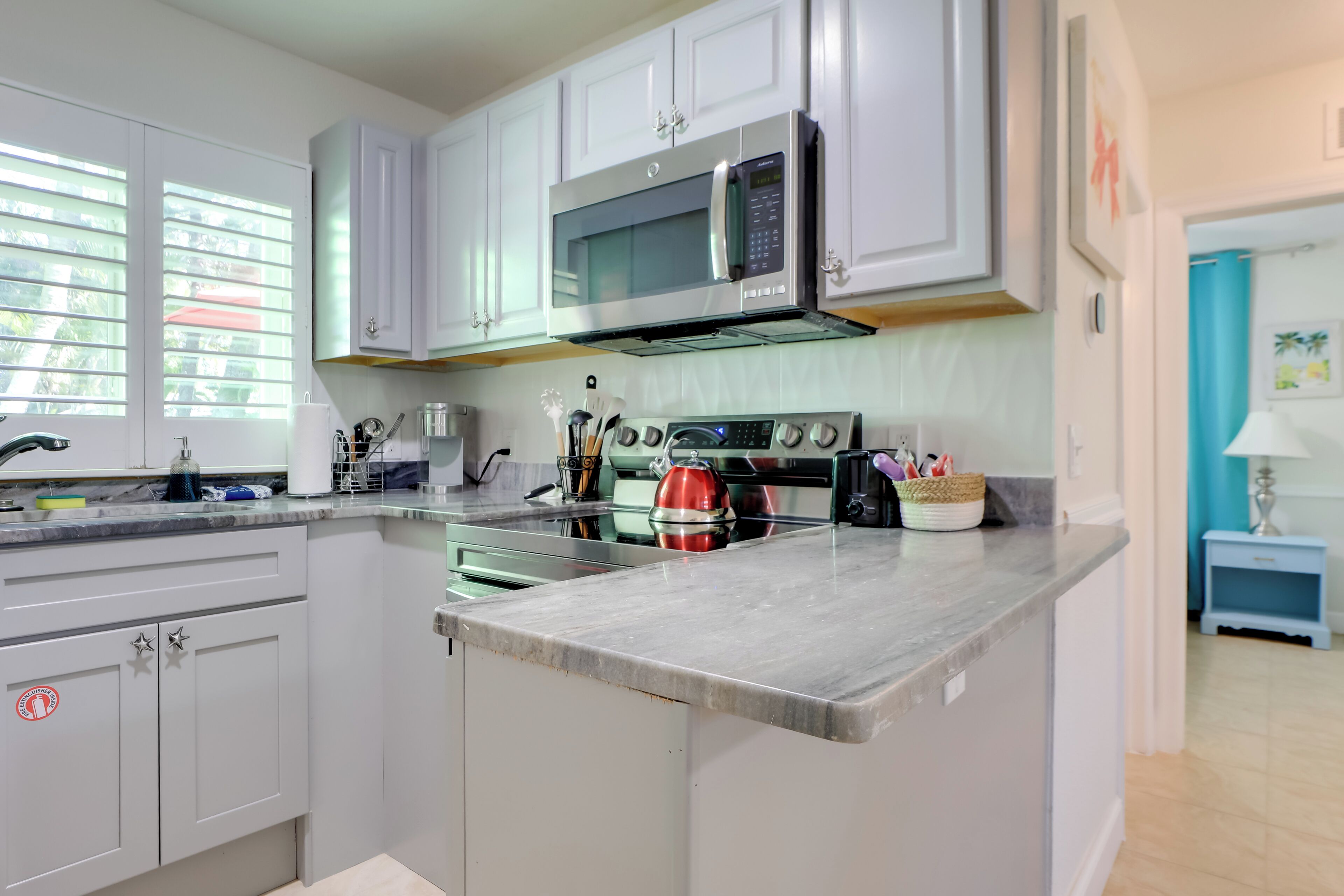 Una cocina moderna con armarios blancos, un microondas de acero inoxidable, un hervidor rojo y una encimera con diversos utensilios de cocina.